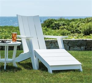 A White Contemporary Adirondack Chair with a Footstool and side table on the grass near a stone wall.